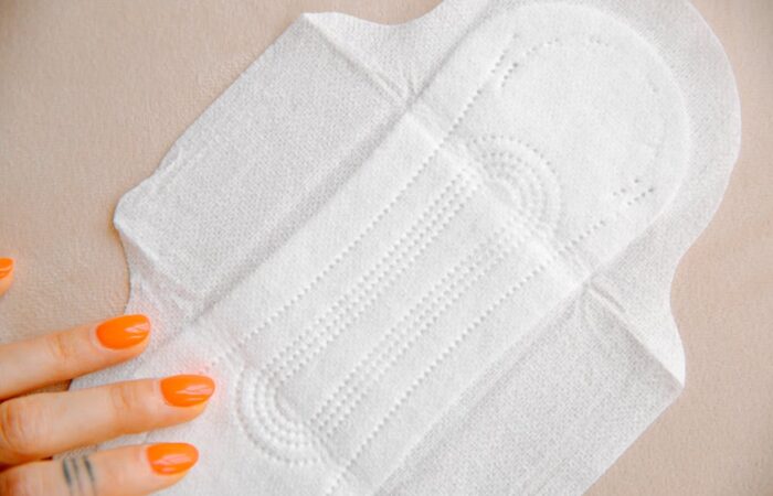 Organic pads in the UAE; A close-up of a white sanitary pad on a light surface, with a hand featuring bright orange nails gently resting beside it.